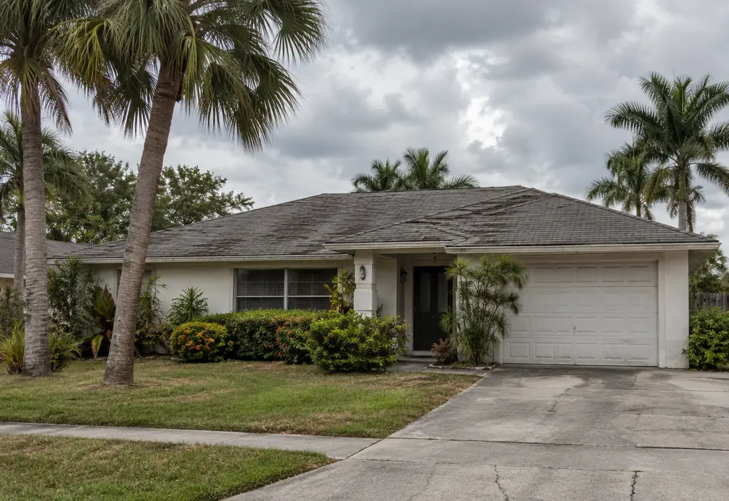An older Florida home with a faded, aged asphalt shingle roof showing curling edges, palm trees, overcast sky, as seen from the street