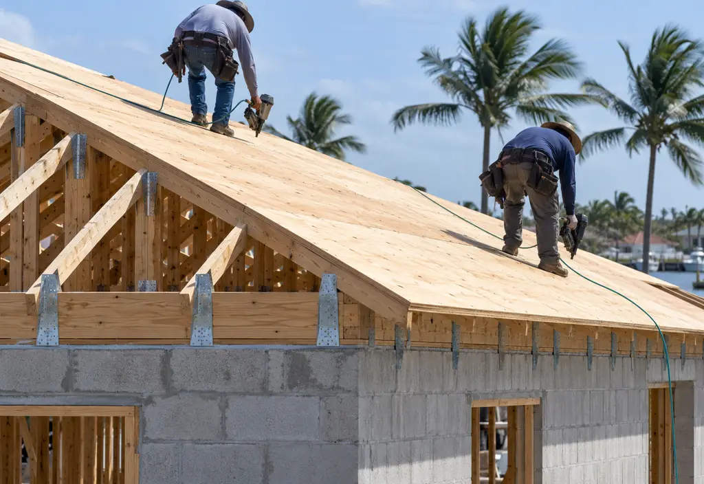 Roofers fastening plywood roof decking with ring-shank nails on a Florida house before underlayment goes on, hurricane straps visible at the wall top plate. Two workers at a distance, faces not visible