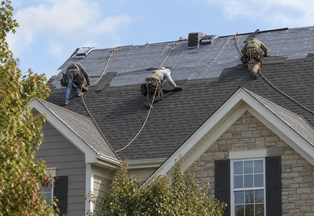 A roofing crew of three workers in plain work clothes and harnesses installing shingles on a pitched roof, seen from the street at a distance, faces not visible, safety ropes attached