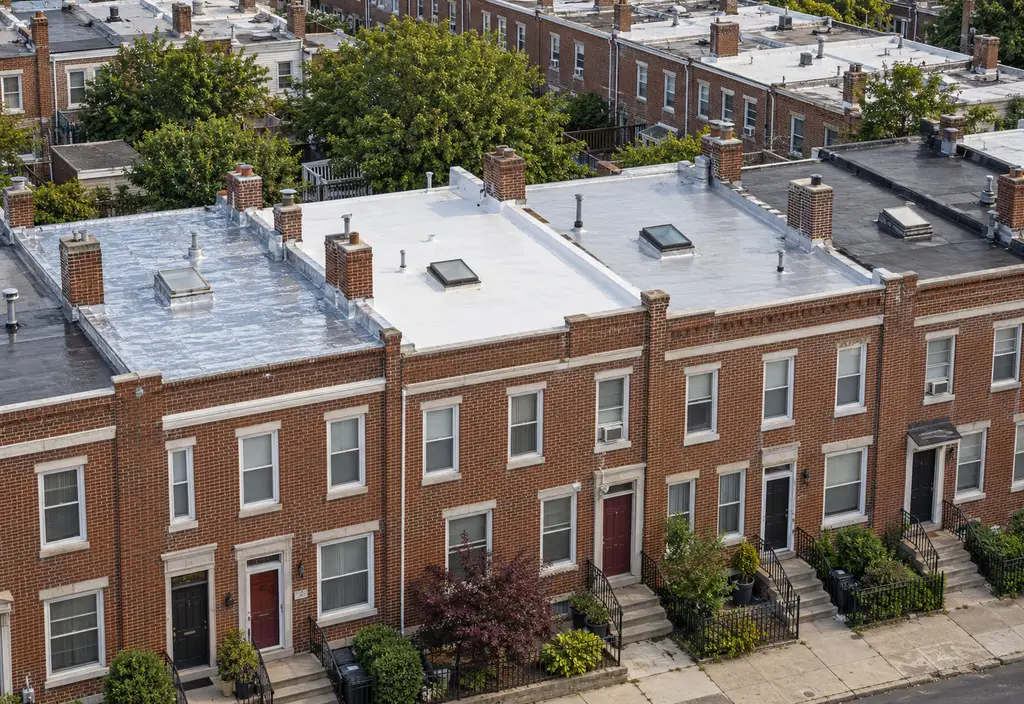 Aerial view of a row of two-story brick rowhouses in Philadelphia with flat roofs behind front parapets, some roofs silver-coated, some new white membrane, chimneys and roof hatches